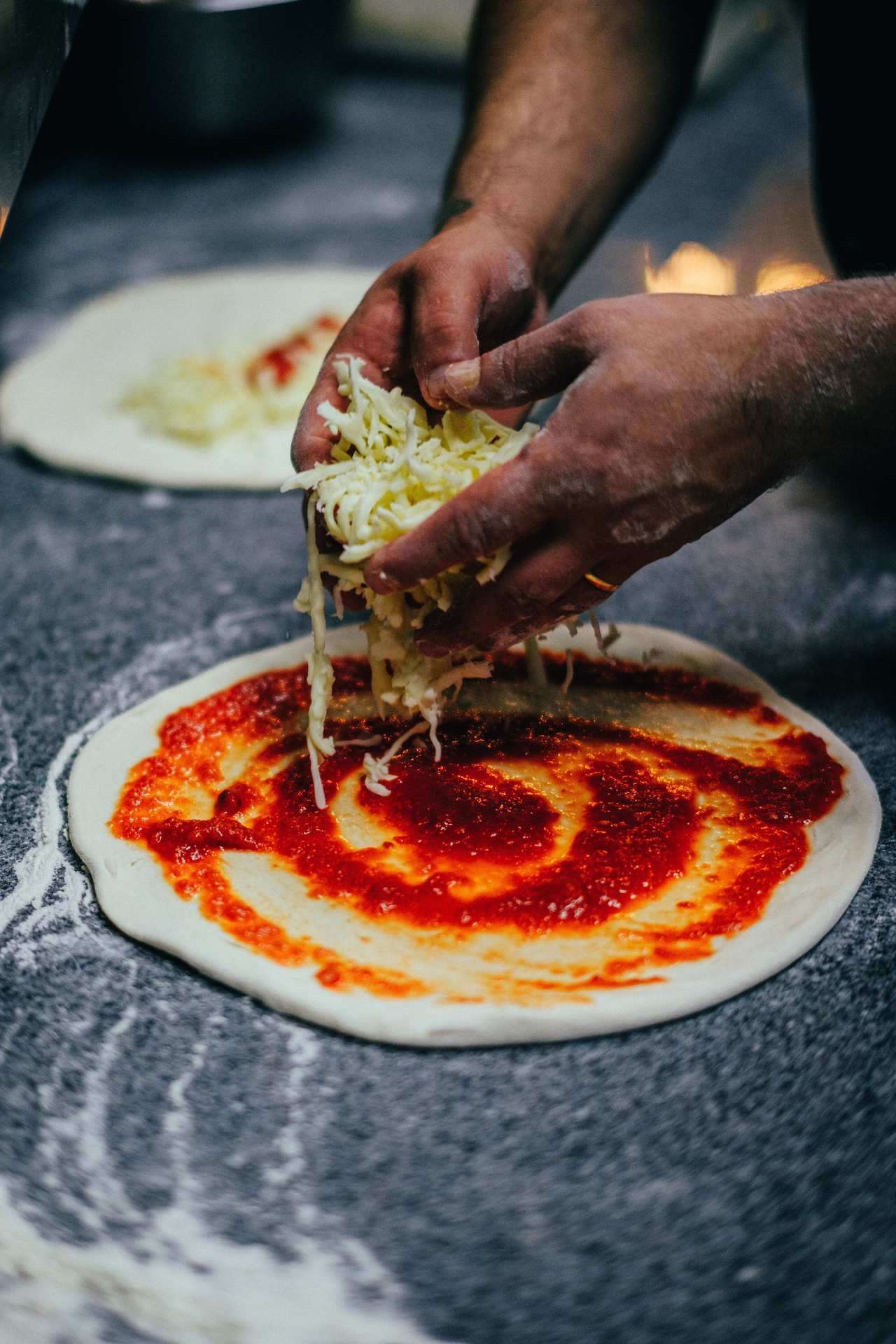 a view of hands spreading cheese on pizza dough covered with tomato sauce