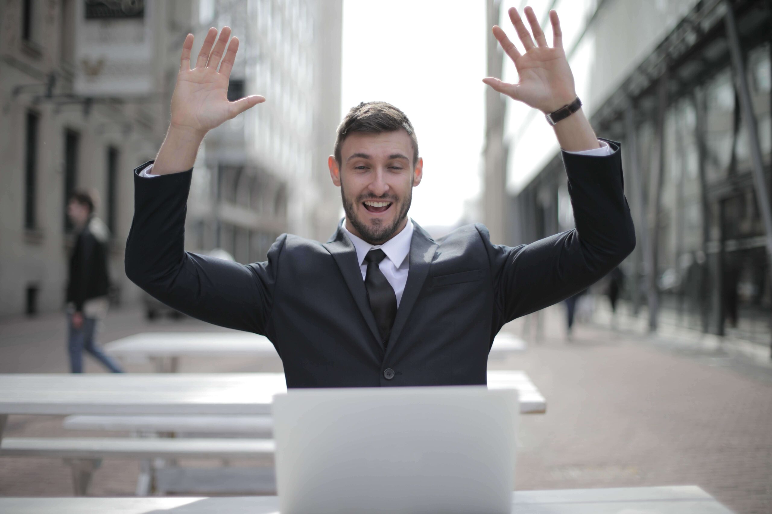 young man in business suit in front of an open laptop raising his hands in triumph