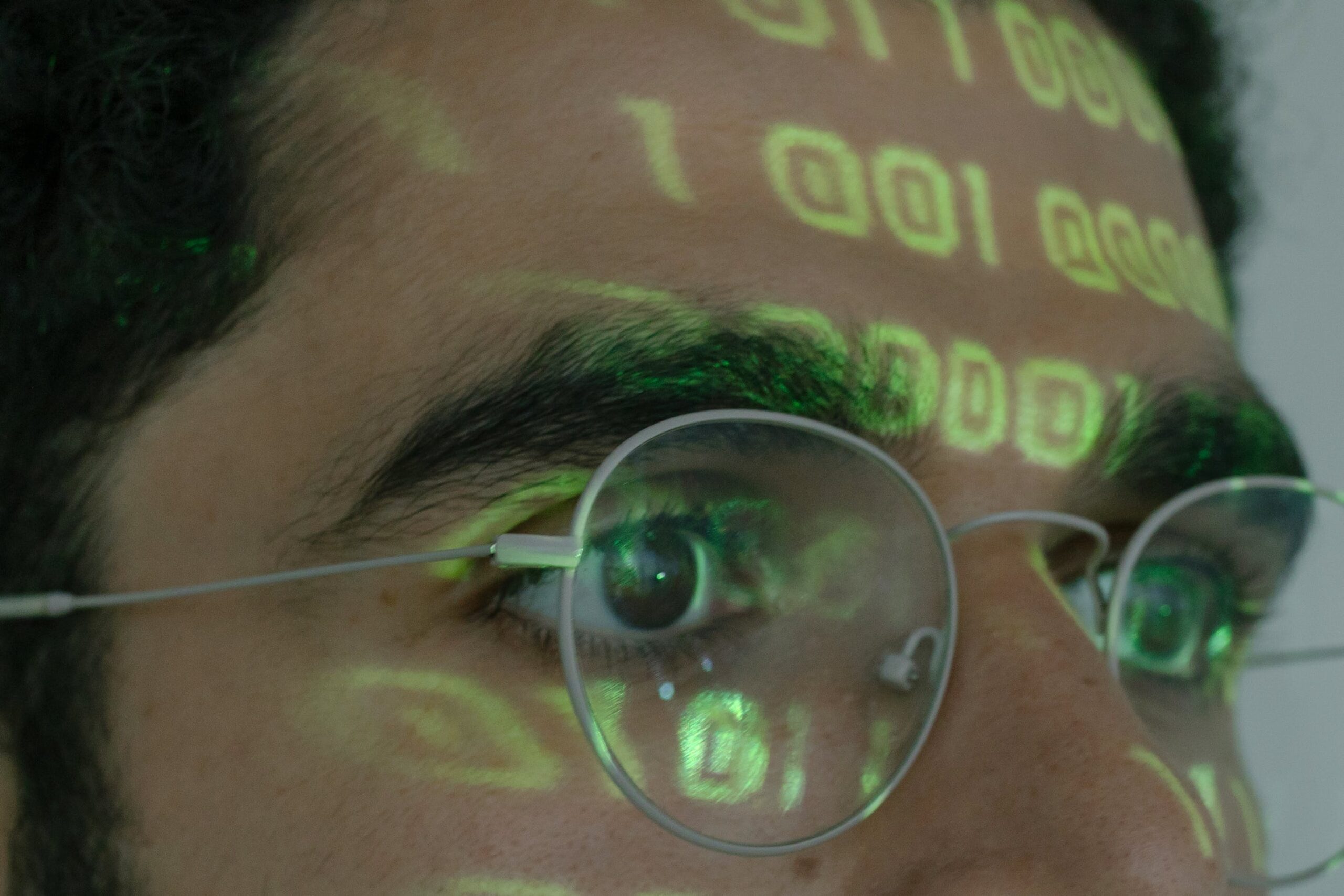 A close up of the face of a dark haired man in glasses with green computer code shining on his face