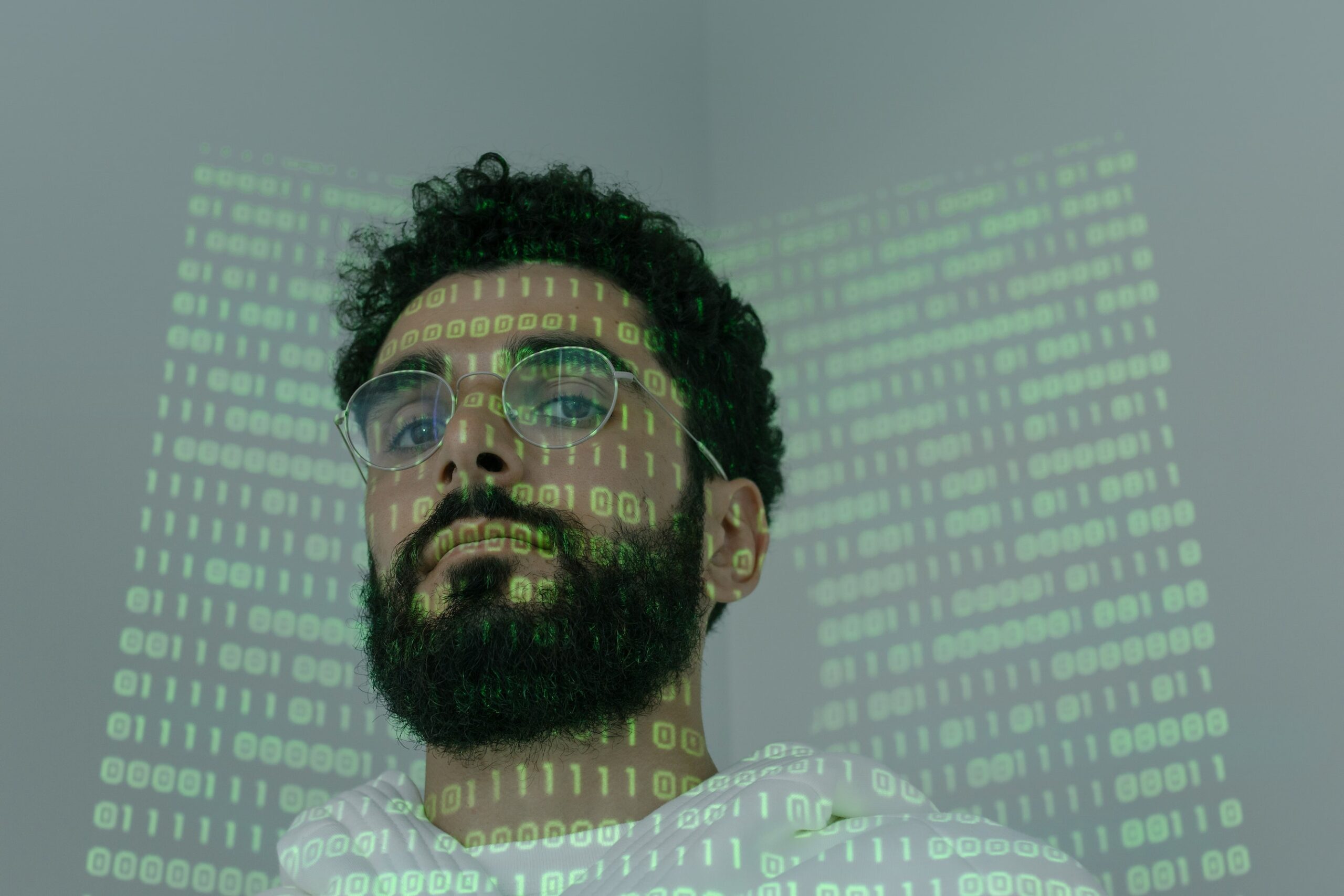 bearded, dark haired man in front of a screen with computer code, part of the code is projected on his face and head