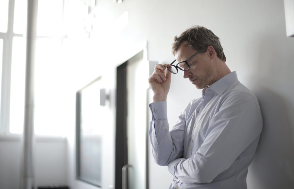 Decorative. A middle aged man takes off his glasses while leaning against a white wall.
