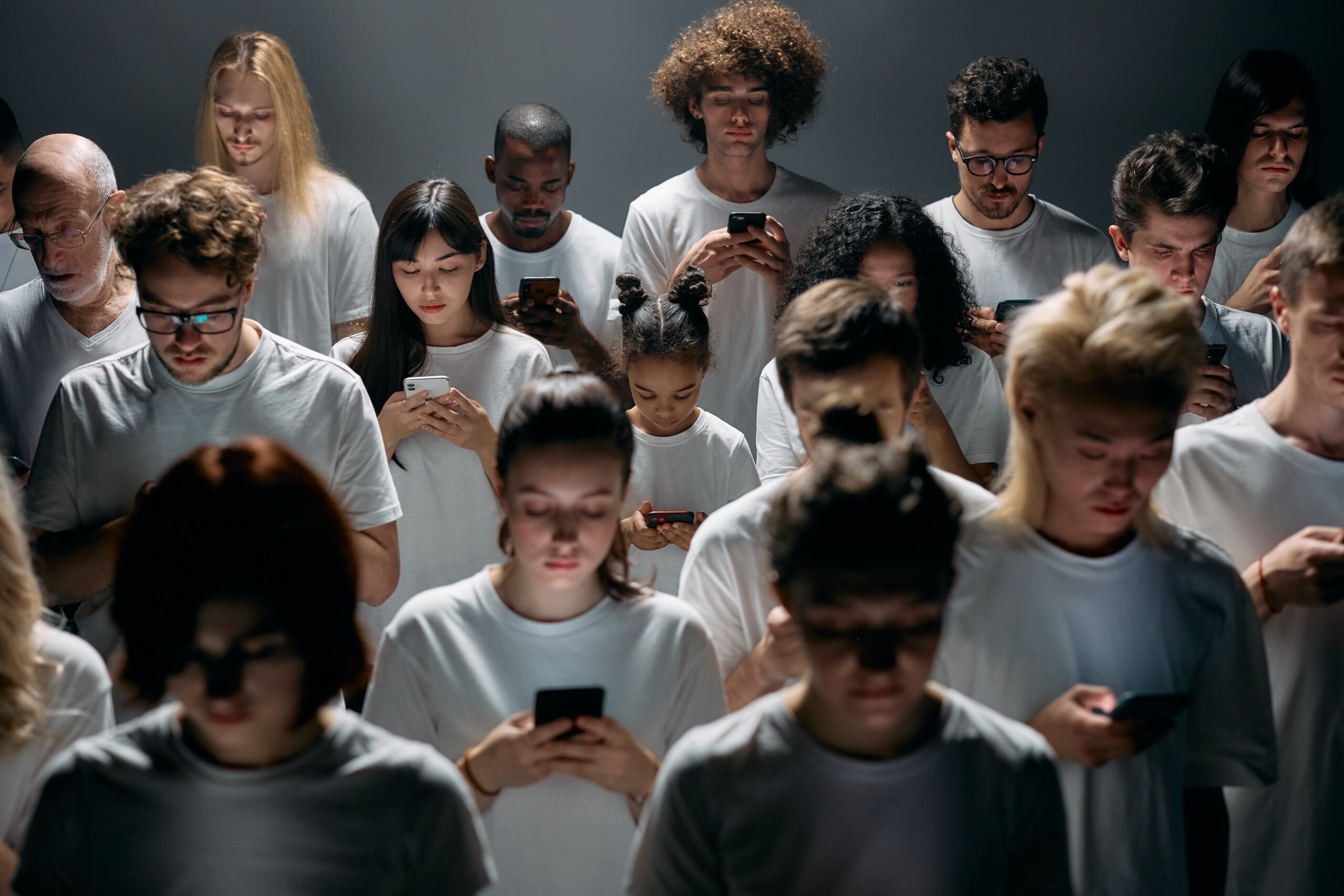 Decorative. A group of people stand staring at their phones in identical clothes. The light is dark everyone except for the blue light on their face.