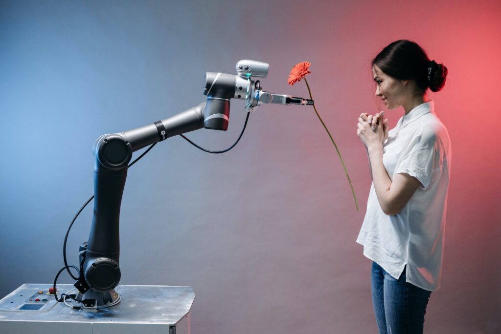 Robot arm against a pale red blue background tenderly offering a single red flower to a young dark haired woman in a white shirt and jeans