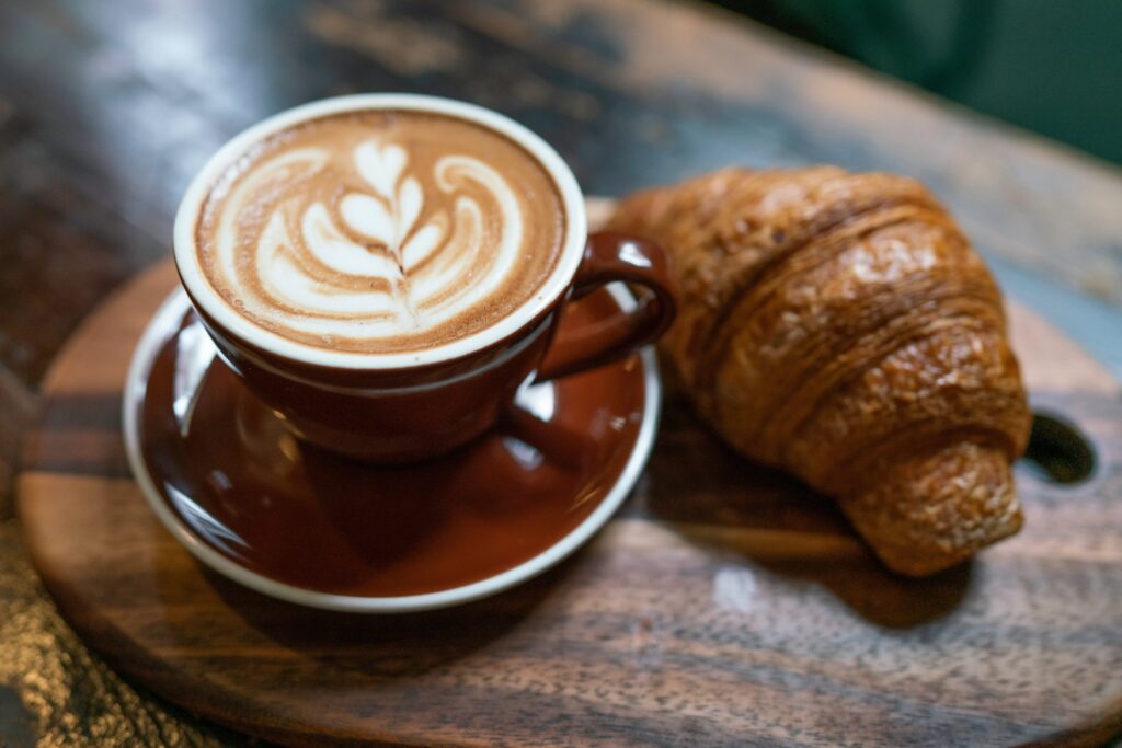 a brown coffee cup on a matching saucer on a wooden table with a plain croissant next to it 