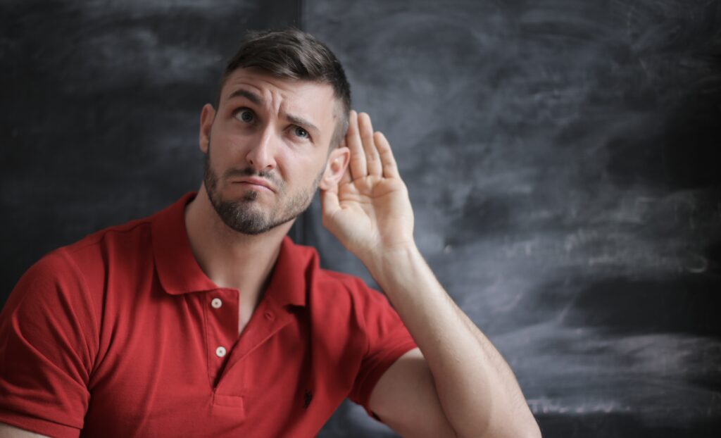 A young man in a red shirt quizzically holding his hand up to his ear