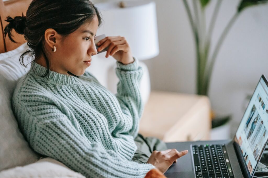 A young, dark haired woman in a light blue sweater sitting on a couch and looking pensively at her laptop