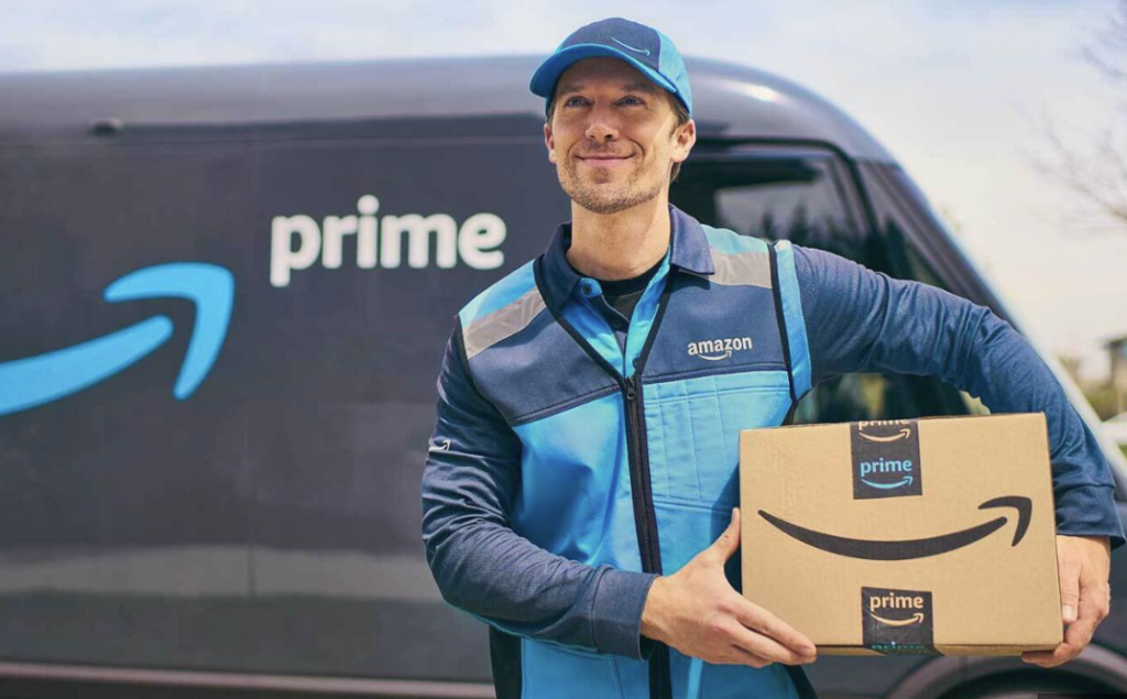 An Amazon delivery man in an Amazon uniform holding an Amazon box in front of a grey Amazon delivery van 