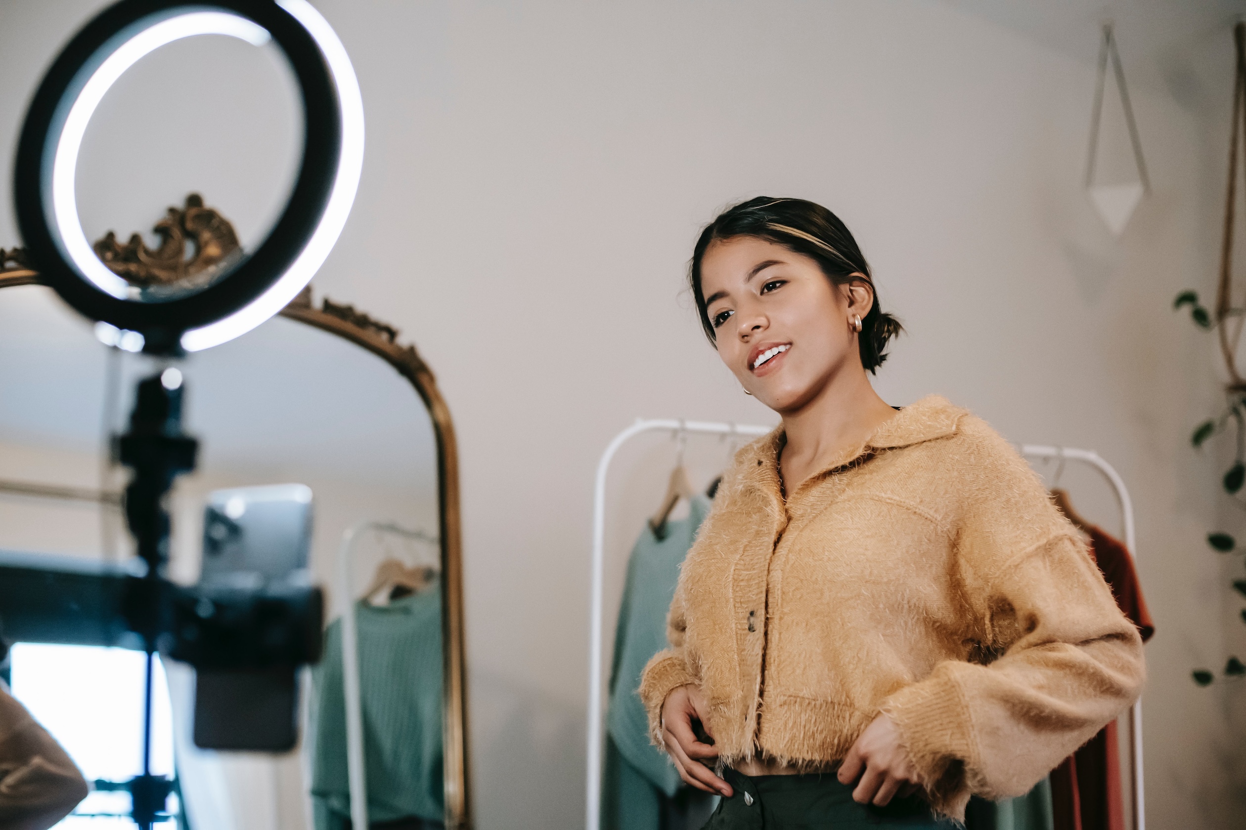 A dark haired young woman modeling a brown sweater while conducting a live stream in front of a mirror and a round ring light