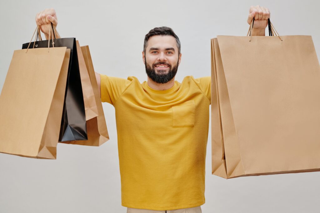 A dark haired, bearded man in a yellow shirt holding up four brown paper shopping bags 