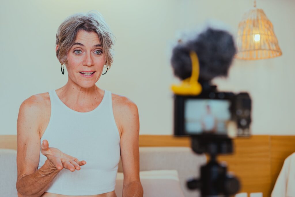 A middle aged light haired woman in a white tank top is talking expressively into her stand held smart phone while recording a video
