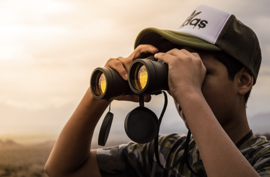 A young man in a baseball cap looking through binoculars across an open field 
