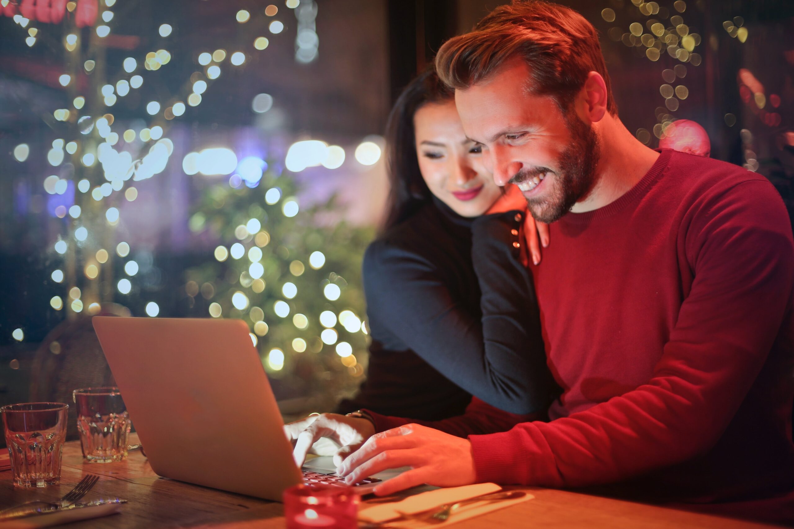 A man and a woman dressed in holiday clothing in front of a christmas tree looking at an open laptop computer