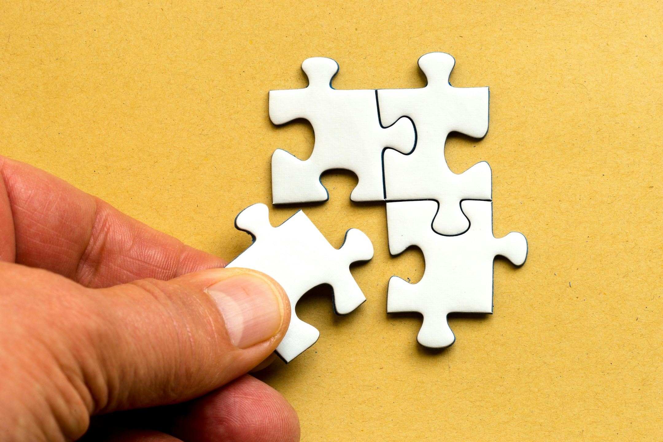 Close up of a hand placing a metallic jigsaw puzzle piece in a group of 4 pieces against a yellow background