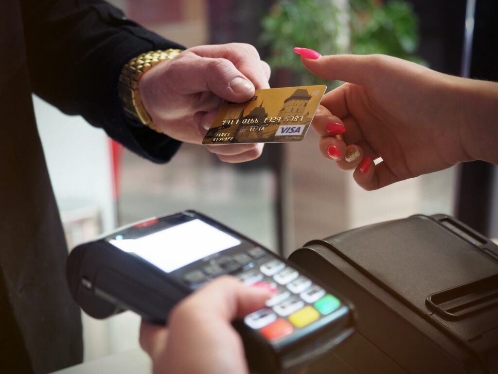 Someone wearing a suit jacket and a gold watch handing a gold Visa card to a merchant using a handheld card reader