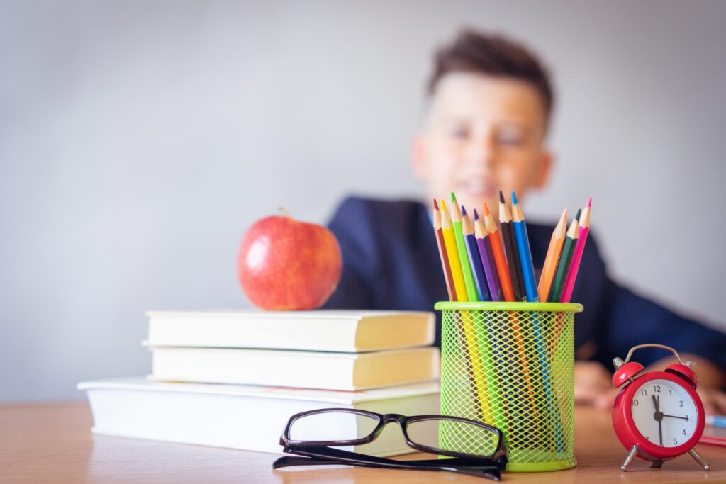 A blurred image of a young man behind a table piled with school supplies including bllks, an apple, pencils, eyeglasses and an alarm clock
