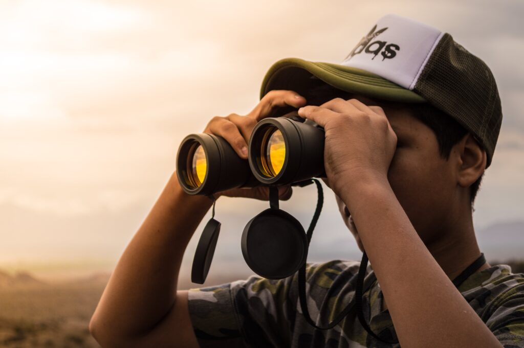 A young man wearing a logo baseball cap is looking through binoculars into the distance 