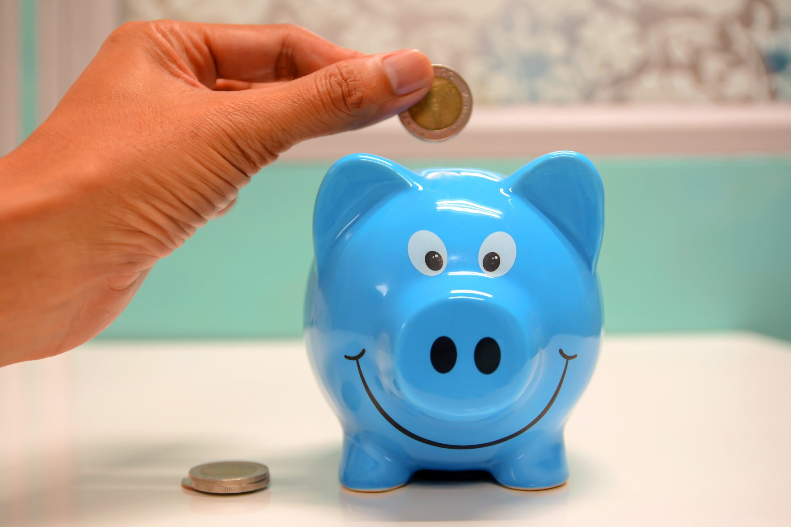 A bright blue piggie bank on a tabletop with a left hand placing a coin in the opening