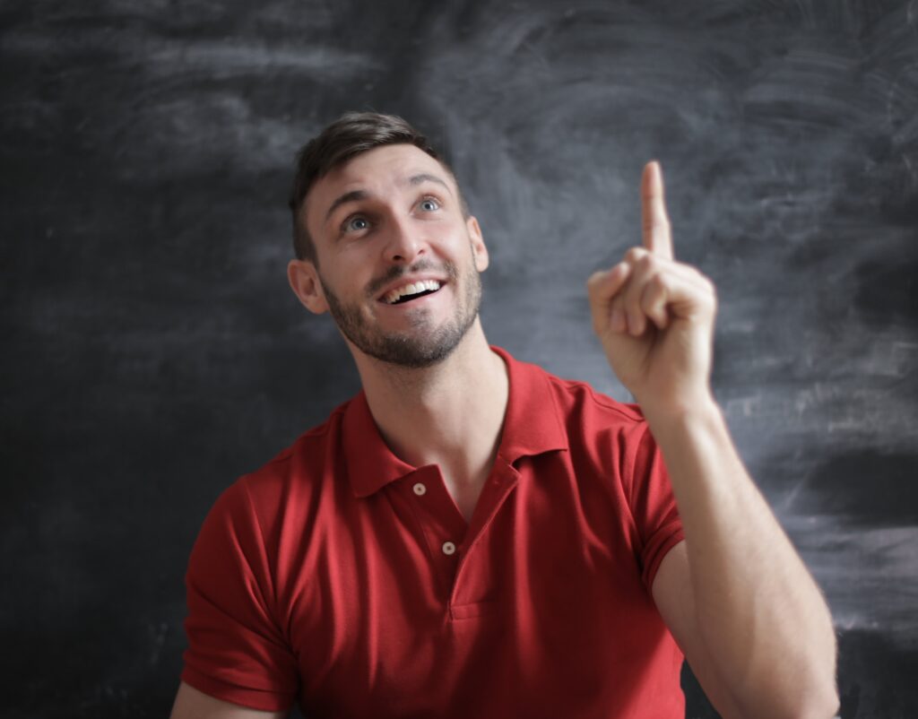 A bearded young man in a red button down short sleeve shirt looking up with a raised hand and index finger 