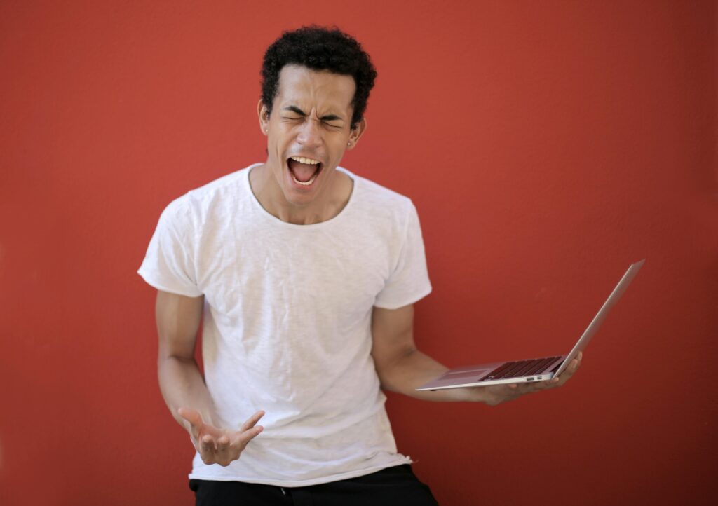 A young, dark haired man wearing a white t shirt screaming and holding an open laptop with his eyes closed 