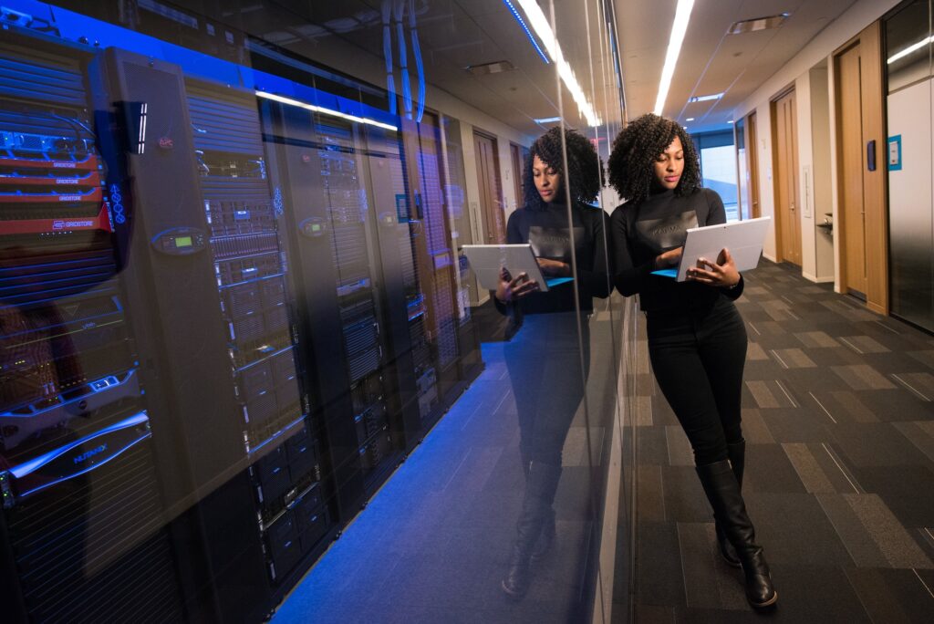 A modern computer server farm behind a glass floor to ceiling window with a young dark haired woman holding a laptop leaning against it 