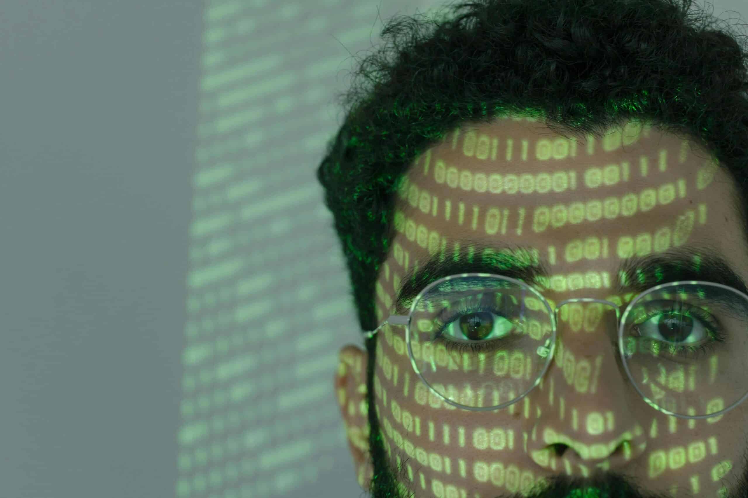 A dark haired man with a beard and glasses looking out into space with green computer code shining on his face