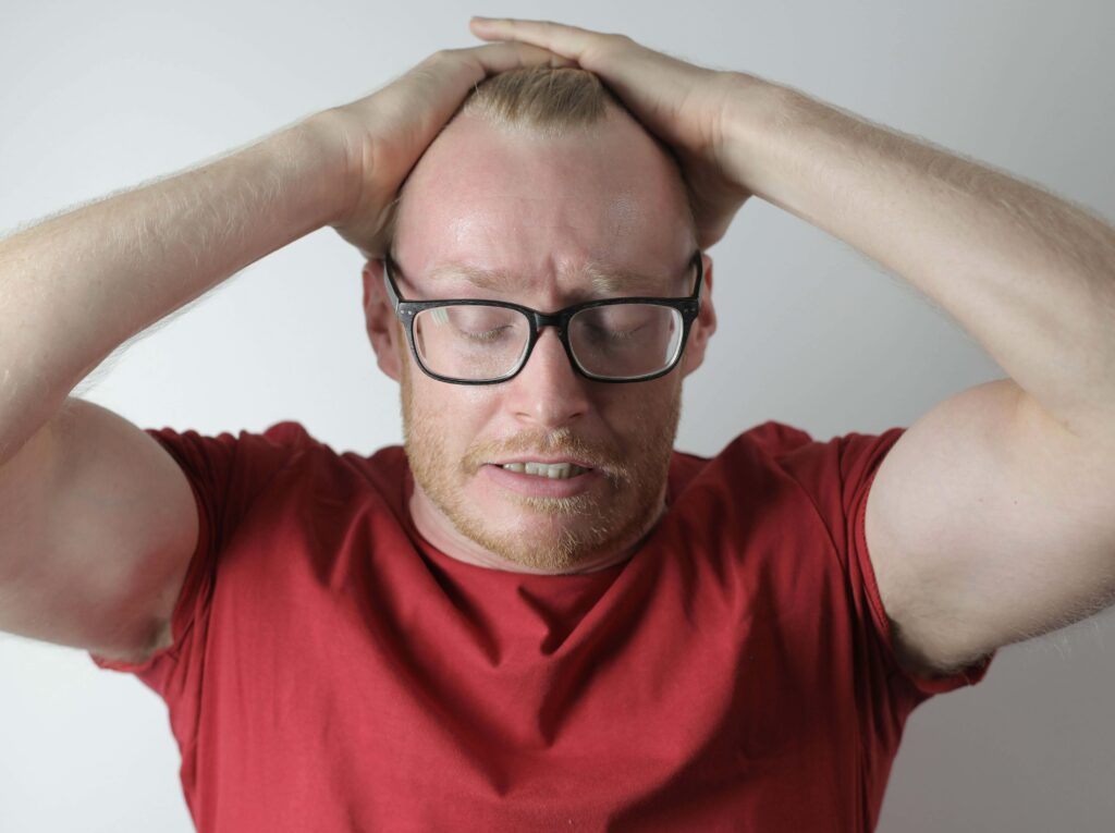 A blond haired man in glasses and a red shirt with his hands on his head and eyes closed in frustration